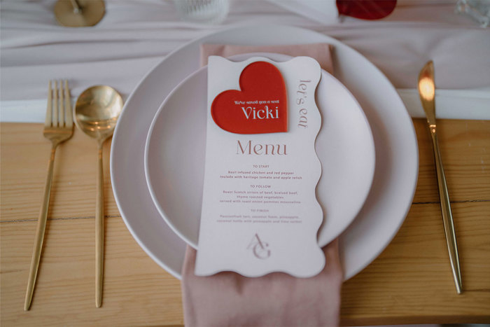 A menu sitting on a plate on a table with a pale pink napkin underneath the plate and a red plastic heart sitting on top of the menu