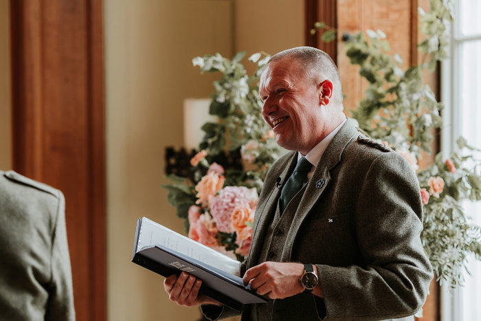 man in a green wool suit smiles as he reads from a blue folder with a nice green and pink display of flowers behind him