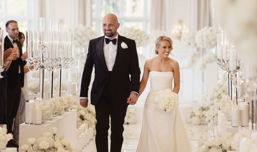 a bride wearing a strapless wedding dress and veil holding hands with a groom wearing a black tuxedo suit in a room filled with white flowers and candles in glass hurricane vases after their wedding ceremony at Trump Turnberry