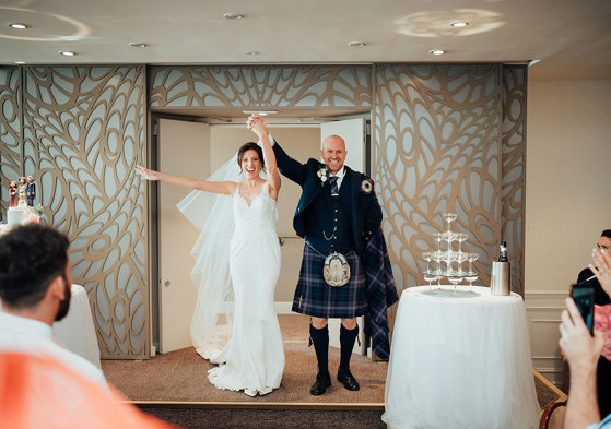 a cheering bride and groom with hands in air walk through a door on an ornate wood-panelled background at seamill hydro