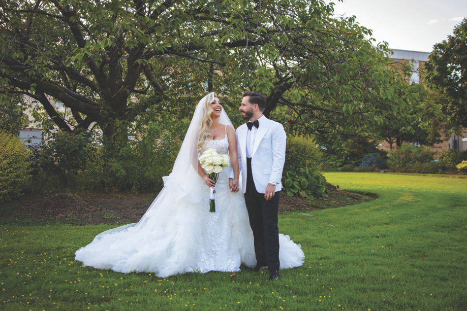 bride and groom wearing white dress and white and black tux standing smiling in grassy park