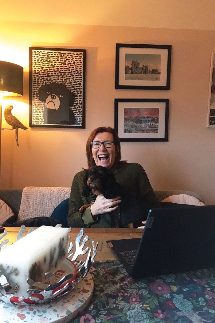 a smiling person holding a dog. They are seated at a table with a laptop in front of them.