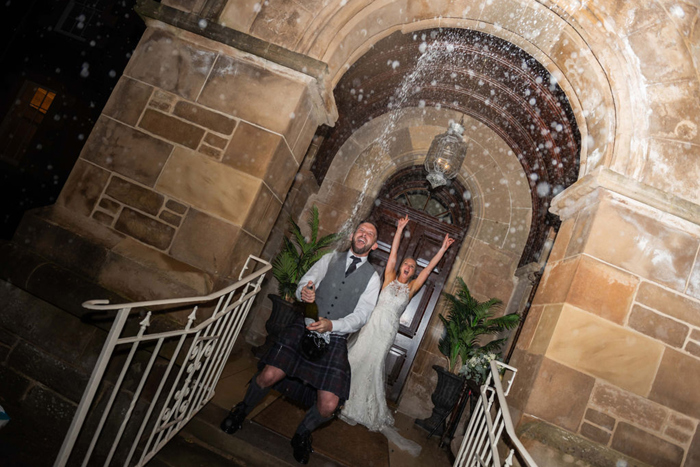 A Person Doing A Champagne Spray Outside A Stone Archway As Another Raises Their Arms Cheering