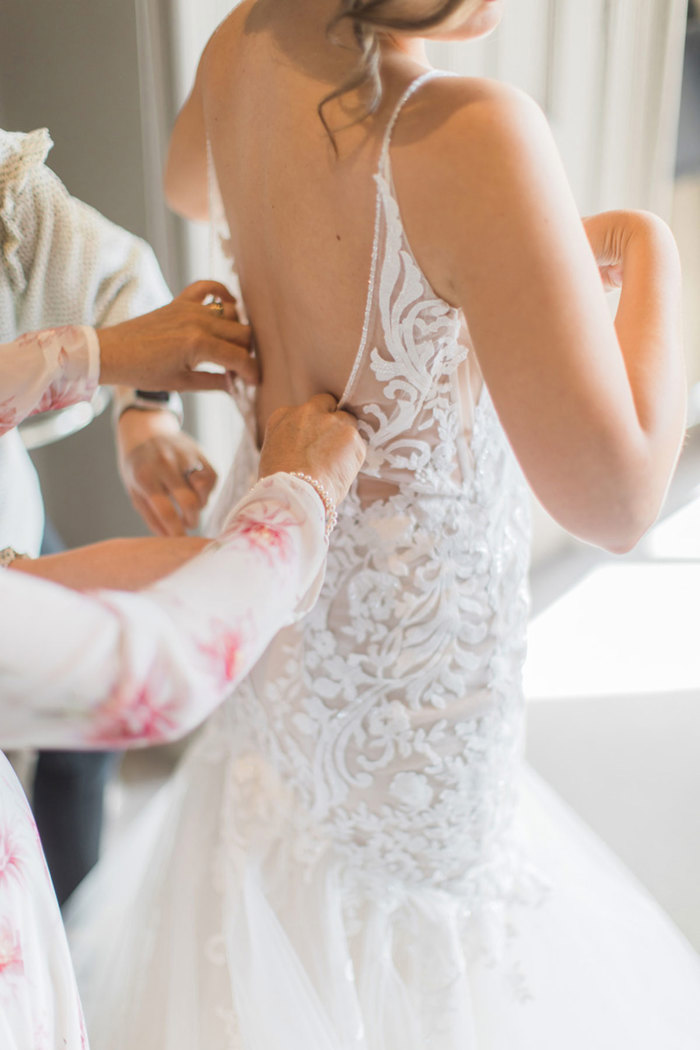 view from back of bride in low-back wedding dress as a loved one with pink and white floral sleeves is carefully buttoning up dress