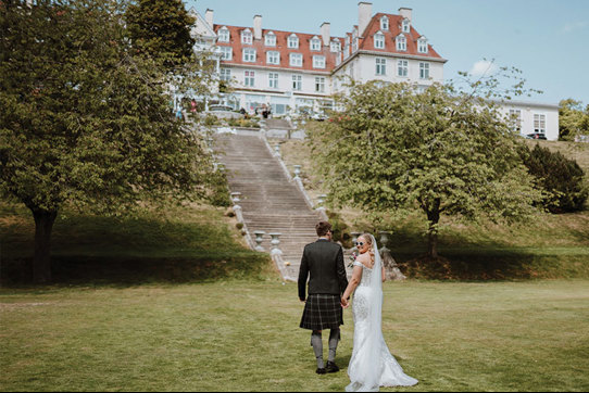 A groom in a kilt and a bride in a white dress stand on a grass lawn at the bottom of a tall set of stone stairs leading up to a large white building with an orange roof