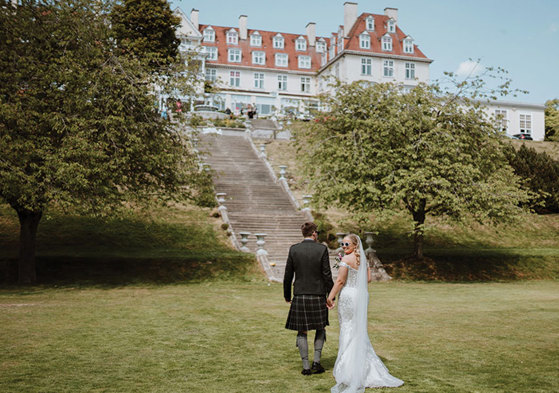 A groom in a kilt and a bride in a white dress stand on a grass lawn at the bottom of a tall set of stone stairs leading up to a large white building with an orange roof