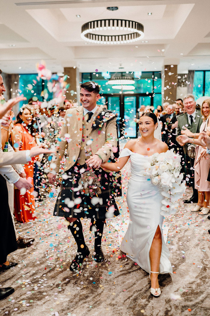 bride in strapless wedding dress with off the shoulder straps holds trailing bouquet in one hand and tall groom in beige and dark green kilt outfit in the other as coloured confetti falls around them