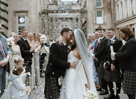 A bride and groom standing outside surrounded by their wedding guests as white confetti is thrown over them