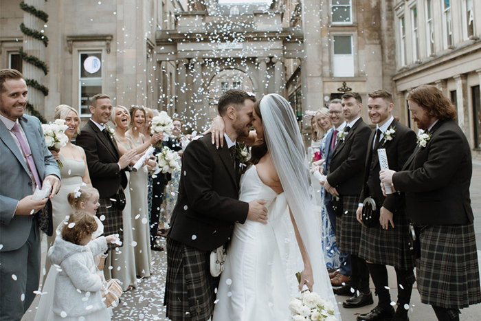 A bride and groom standing outside surrounded by their wedding guests as white confetti is thrown over them