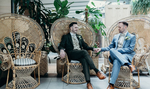 two groom sitting on wicker peacock chairs holding hands