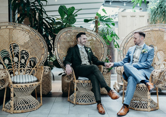 two groom sitting on wicker peacock chairs holding hands