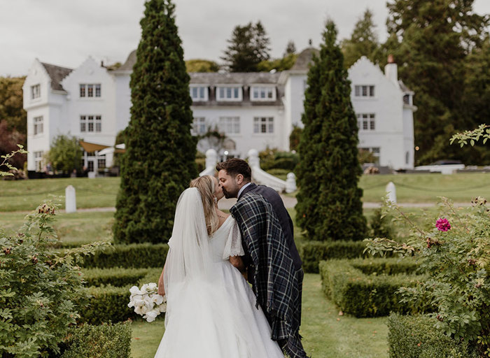 a bride and groom walking hand in hand with back to the camera kiss in the gardens outside Achnagairn Castle, the white facade of the castle visible in the background