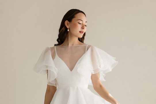 dark haired model wears v-neck white wedding dress with sheer layer over bodice providing fluttery sleeves