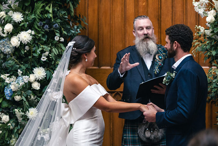 celebrant Andrew D Scott standing against a large wooden door thats surrounded by blue and white flower decorations leads a wedding ceremony as a bride and groom hold hands and look at him pensively