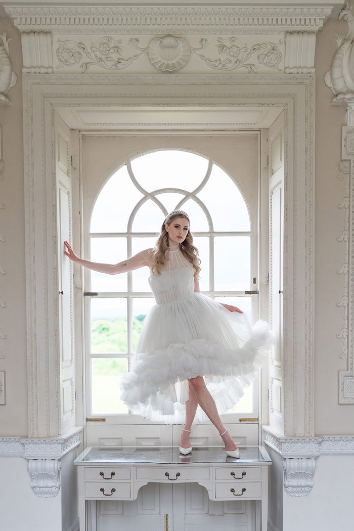 woman stands on top of dressing table in windowsill while wearing a white tulle dress that's short in the front and more midi long in the back