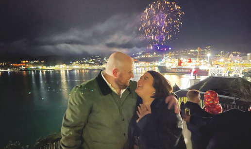 man and woman smiling at each other with water, ferry and fireworks in the background