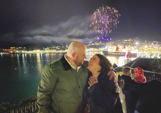 man and woman smiling at each other with water, ferry and fireworks in the background