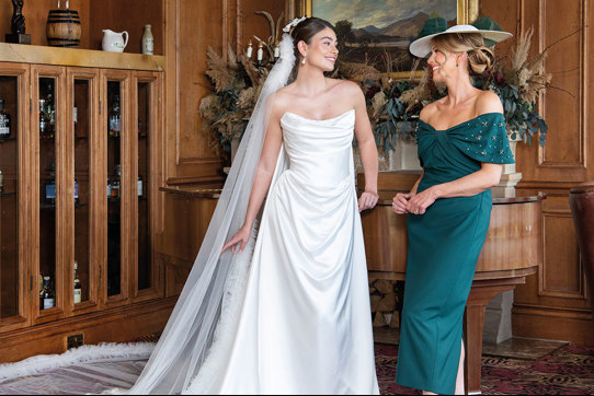 A bride wearing a strapless white wedding dress and long veil and a mother of the bride wearing forest green dress