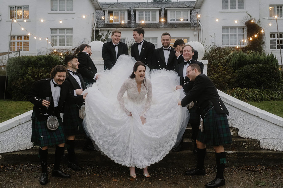 a bride wearing a wedding dress outside Achngairn Castle. She is surrounded by laughing men in kilts holding her skirt.