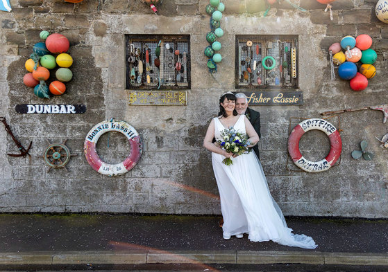 a windswept bride and groom standing in front of a stone building that's adorned with sea paraphernalia.