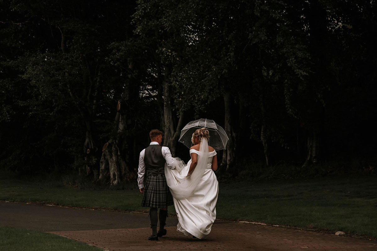 a bride and groom under an umbrella walking through woodland at The Vu countryside wedding venue central Scotland