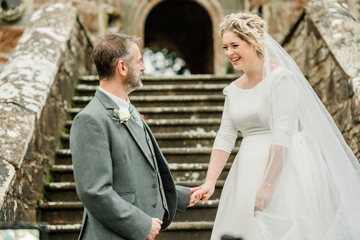 bride in white wedding dress and veil and groom in grey suit jacket stand facing each other, hand in hand on outdoor brick staircase