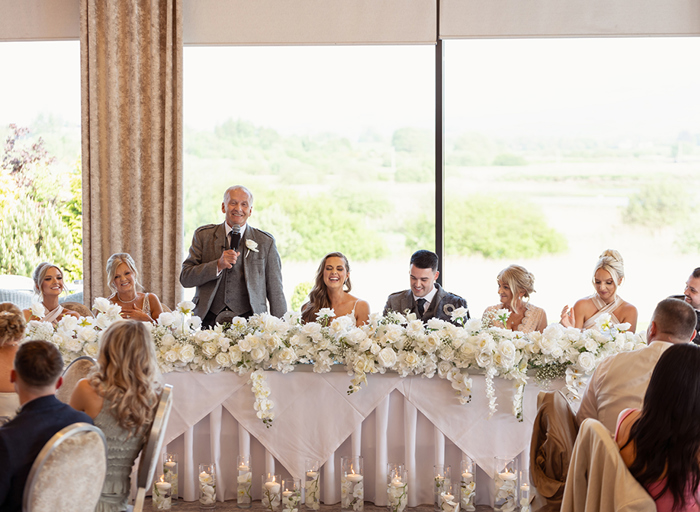 a row of guests and bride and groom seated at a top table at a wedding as a person stands with a microphone