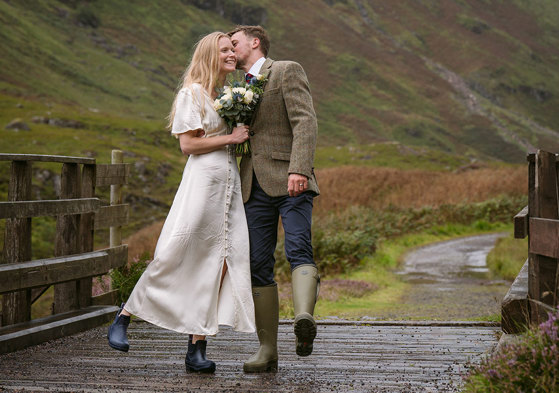 a groom kissing a bride on the cheek. They are both wearing wellies and standing on a wooden bridge in Glencoe