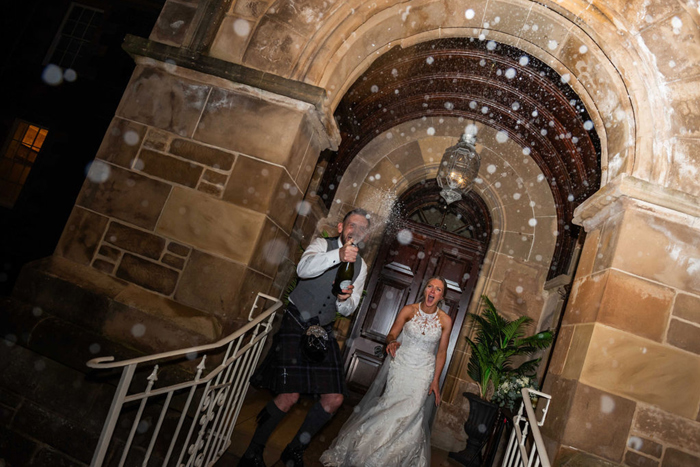 A Person Doing A Champagne Spray Outside A Stone Archway As Another Cheers On