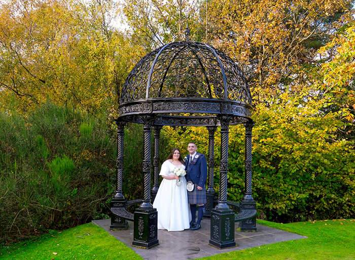 A bride in a wedding dress and a groom in a blue kilt stand under a metal gazebo in a garden