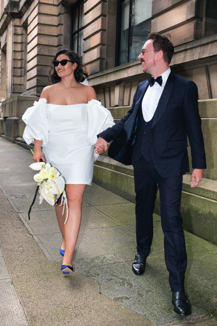 A bride in a short wedding dress, blue high heels and sunglasses holds a white flower bouquet in one hand and her husband's hand in the other, he wears a navy tuxedo and sunglasses