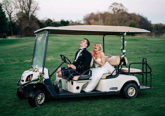 a laughing bride and groom on a golf buggy on a lawn