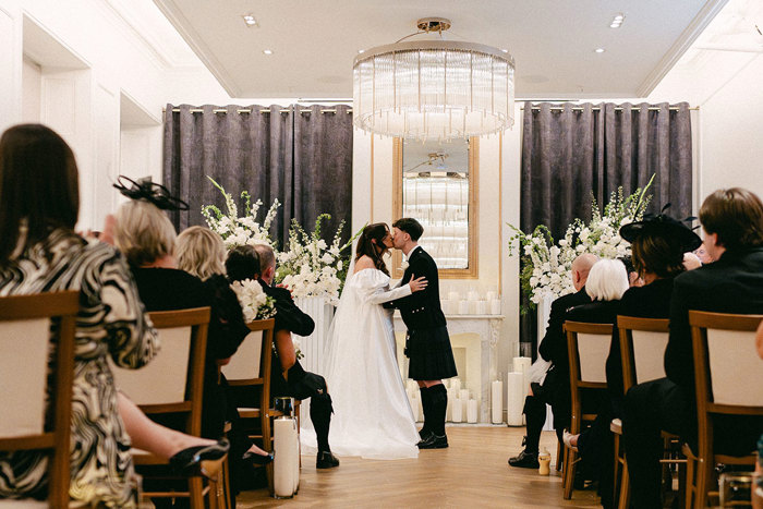 brunette bride in long veil and wedding dress with puff sleeves and groom in full dark tartan kilt outfit kiss at the top of the aisle in a modern ceremony space