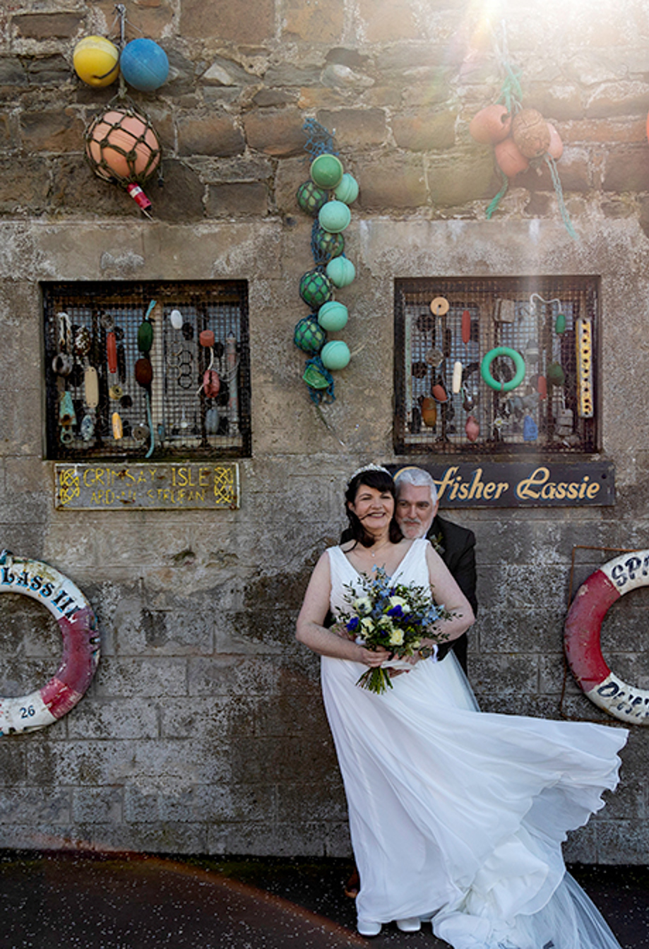 a windswept bride and groom standing in front of a stone building that's adorned with sea paraphernalia