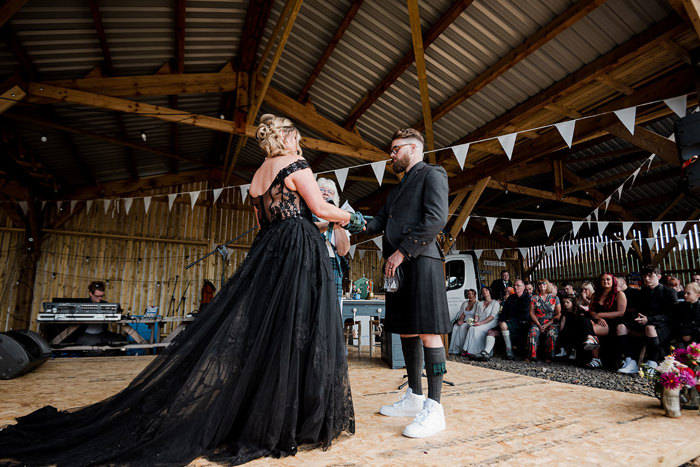 Wedding Ceremony With A Bride And Groom Wearing Black And Guests Seated In Rows Looking On And White Bunting Hanging From Ceiling