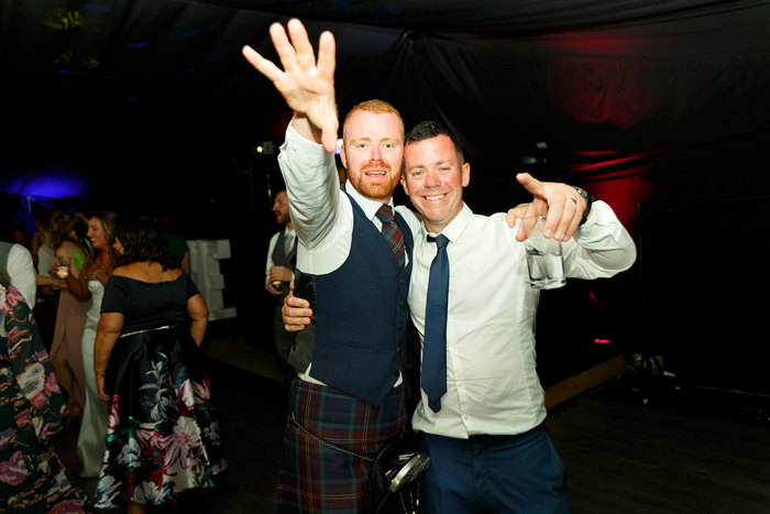 A man in a kilt and a man in a suit look towards the camera and hold their hands up in celebration