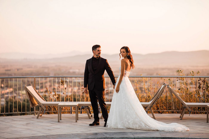 a bride and groom standing in front of a railing with sun loungers and a sunset view behind them.