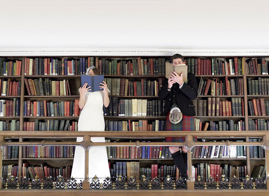 A bride in a wedding dress and a groom in a red and green kilt stand in front of bookshelves of books and hold a book over their faces