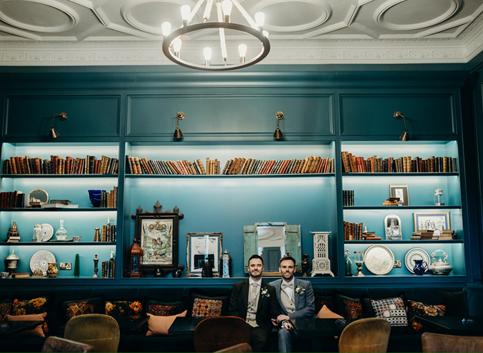 two grooms on their wedding day pose below an illuminated bookcase and cabinet at Kimpton Charlotte Square Hotel in Edinburgh