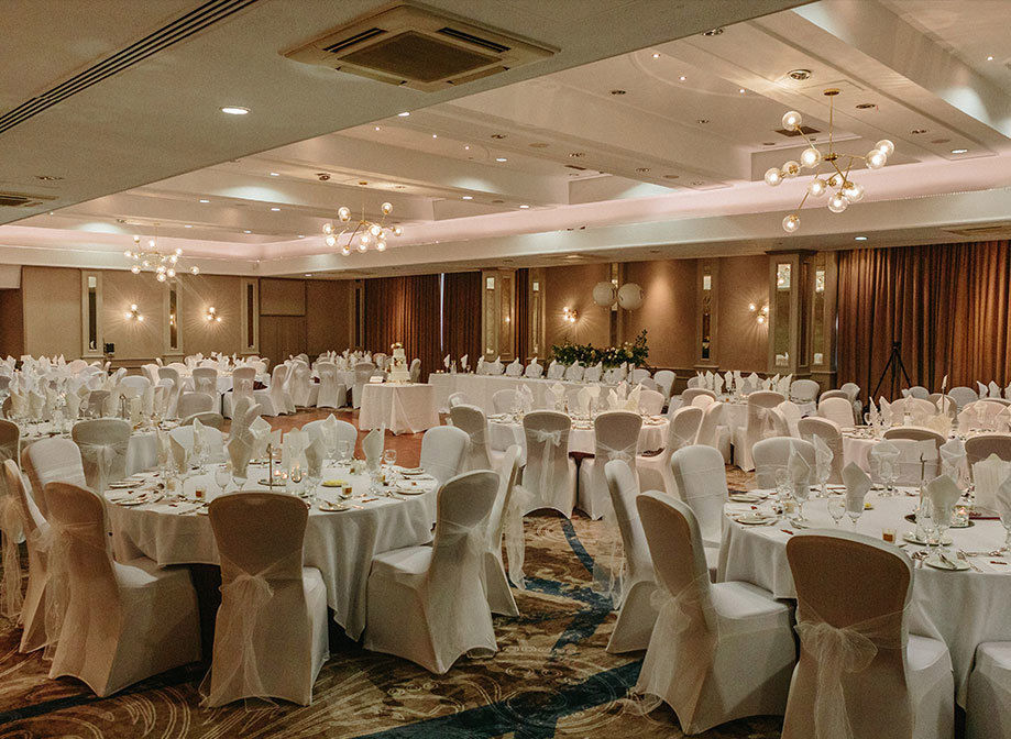 A function room with round tables with white table cloths, chairs with white table covers and white bows and place settings for a meal