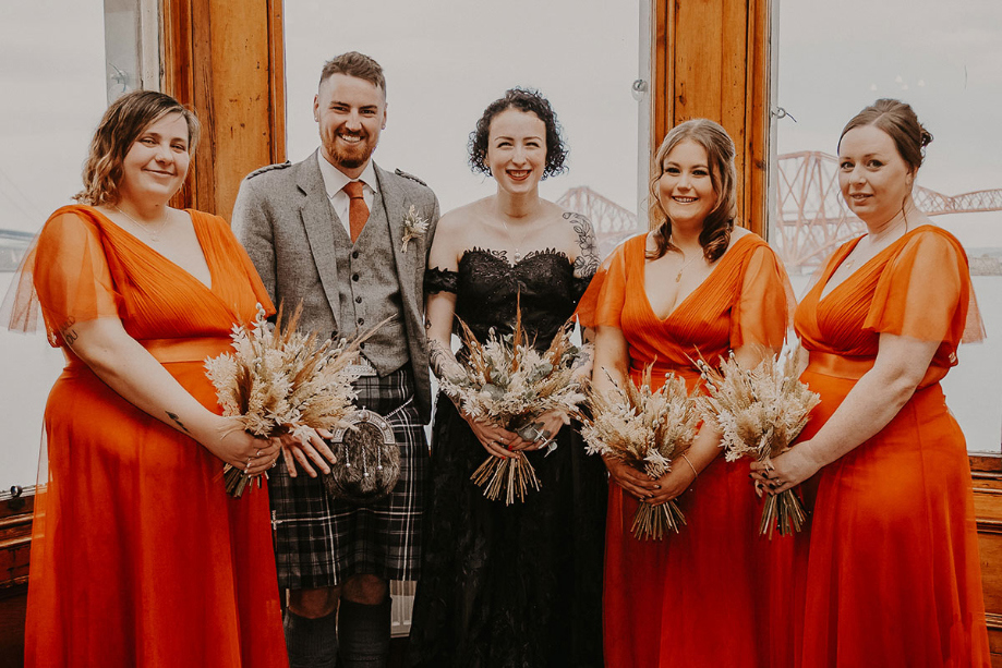 Bride and groom standing with bridesmaids in orange dresses holding dried bouquets 