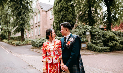 a bride wearing a red and gold Chinese qun kua with a groom wearing a Scottish kilt standing outside a tall Victorian stone building with gardens