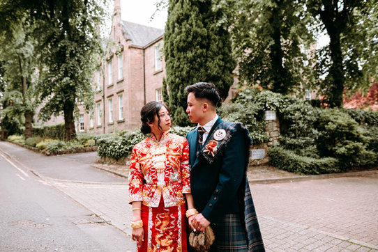 a bride wearing a red and gold Chinese qun kua with a groom wearing a Scottish kilt standing outside a tall Victorian stone building with gardens