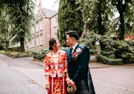 a bride wearing a red and gold Chinese qun kua with a groom wearing a Scottish kilt standing outside a tall Victorian stone building with gardens