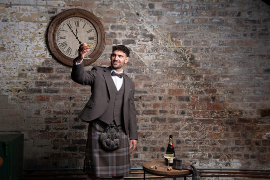 A person wearing a Jura Mist tartan kilt outfit from MacGregor and MacDuff raising a champagne glass. They are standing in front of a rustic brick wall underneath a large clock.