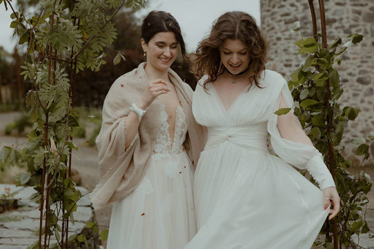 two laughing brides looking downward while standing outside under a foliage-covered wire arch in a garden with old stone building in background