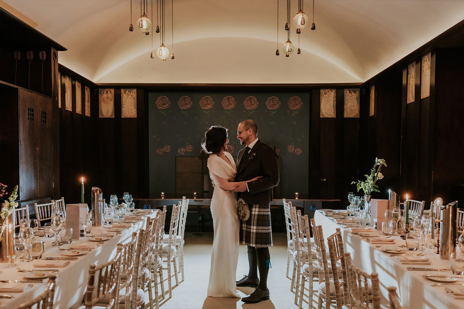A bride in a white dress and a groom inn a kilt stand with their arms around each other next to two long dining tables 
