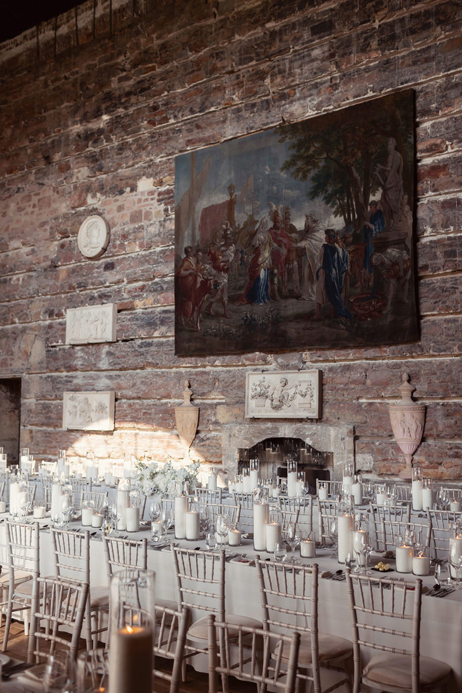 Long tables set for a wedding dinner with chiavari chairs, candles and white flowers. There is an exposed brick wall in the background with large painting.