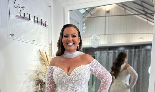 smiling dark-haired model wears off the shoulder long sleeve shimmery wedding dress and a tulle neck scarf