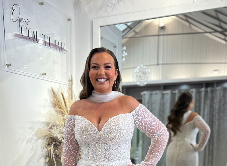 smiling dark-haired model wears off the shoulder long sleeve shimmery wedding dress and a tulle neck scarf